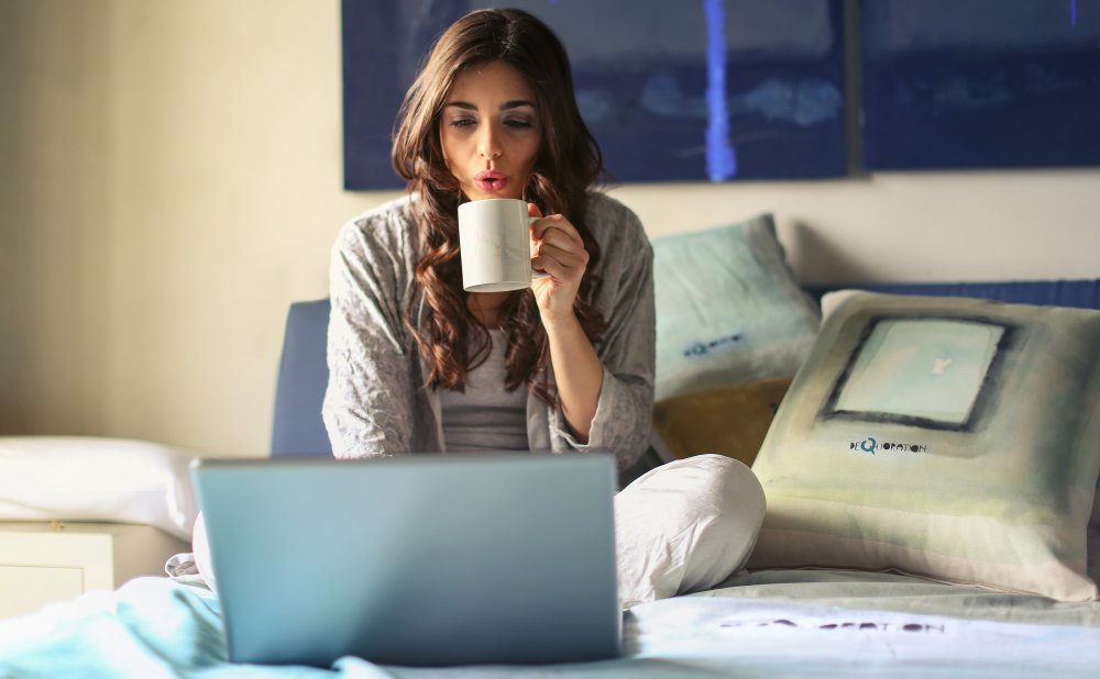 Mujer mirando su ordenador, sobre su cama mientras se toma un café. Lo que simboliza la comodidad de llevar un proceso terapéutico online, desde la comodidad de tu casa.